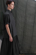 A woman with short brown hair stands in profile, wearing the Work Shirt | Typewriter Cotton Black. She is in front of a dark, semi-transparent fabric backdrop.