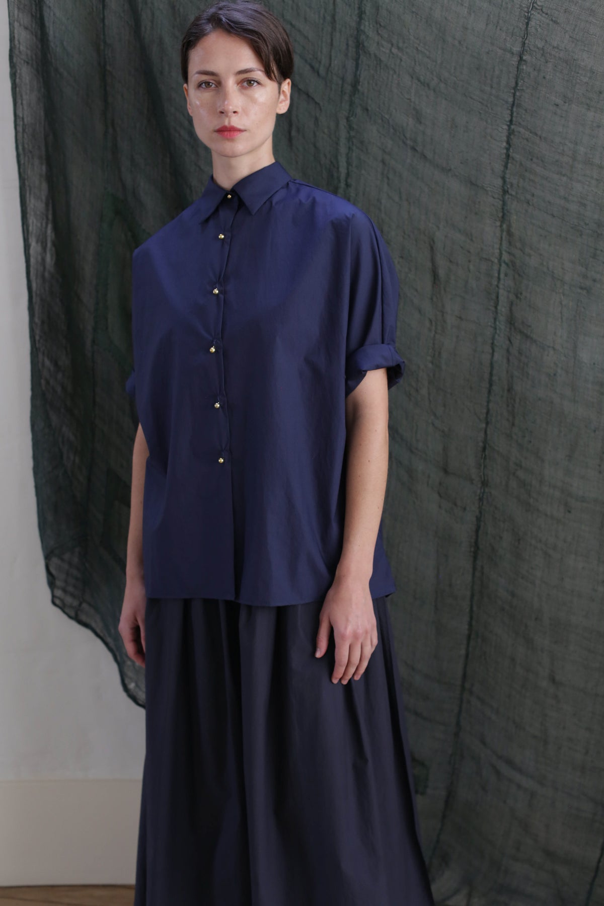 A person indoors wears the Work Shirt | Typewriter Cotton Navy, a loose-fitting navy blue button-up with brass buttons and rolled sleeves, paired with a matching flowing skirt. A textured dark green fabric hangs in the background.