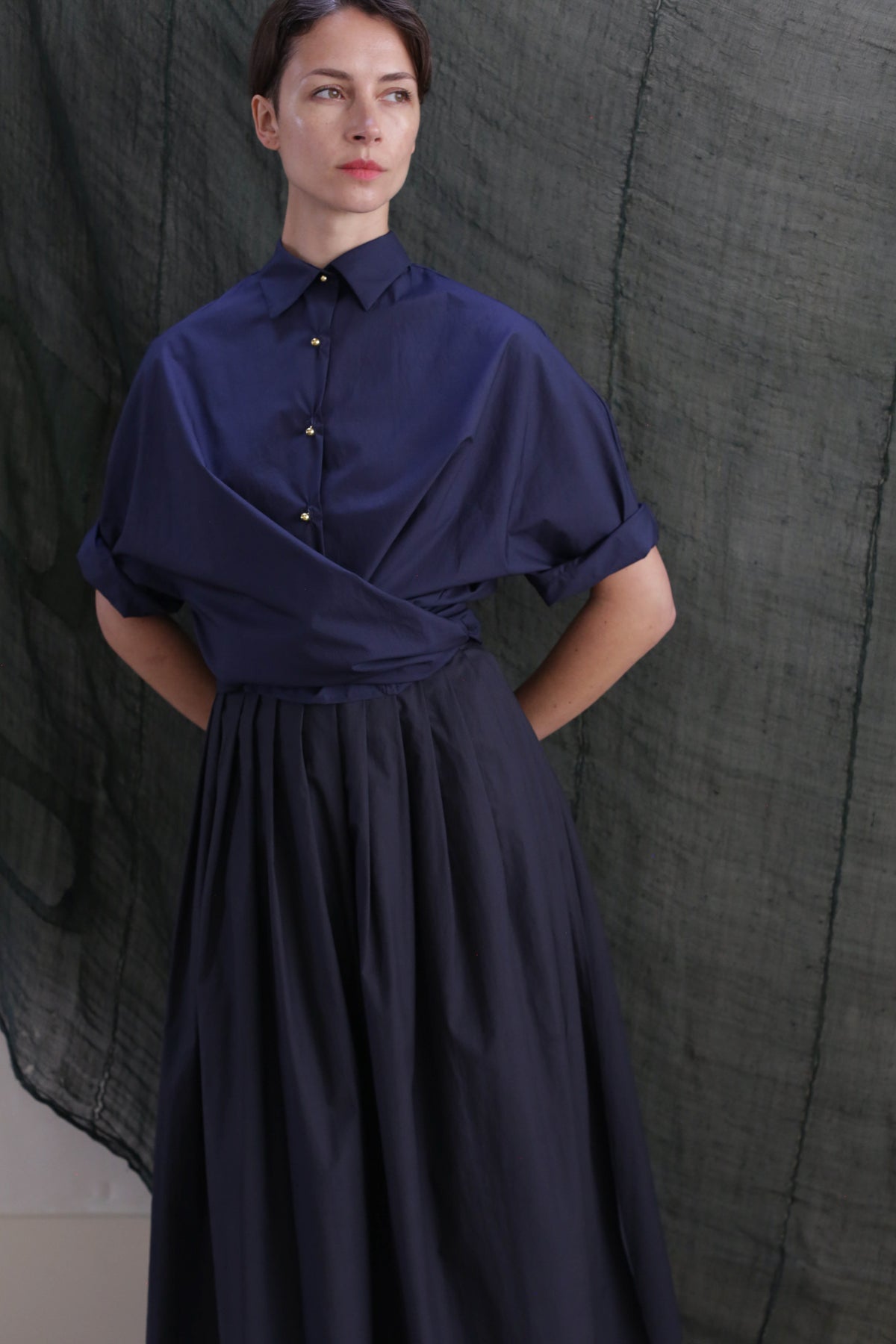 A person wearing the Work Shirt in Typewriter Cotton Navy, featuring brass buttons and short sleeves, stands before a textured dark green fabric backdrop, gazing slightly to the side with hands behind their back.