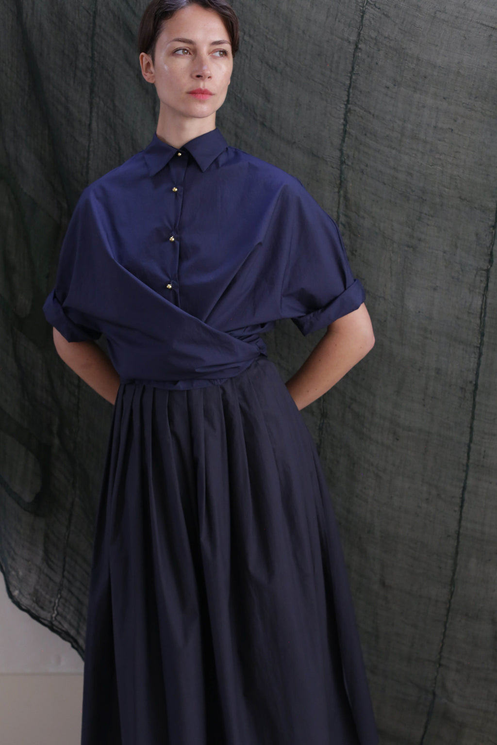 A person wearing the Typewriter Wrap Skirt in navy, featuring a pleated cotton design, stands with hands behind their back against a dark green textured fabric backdrop.