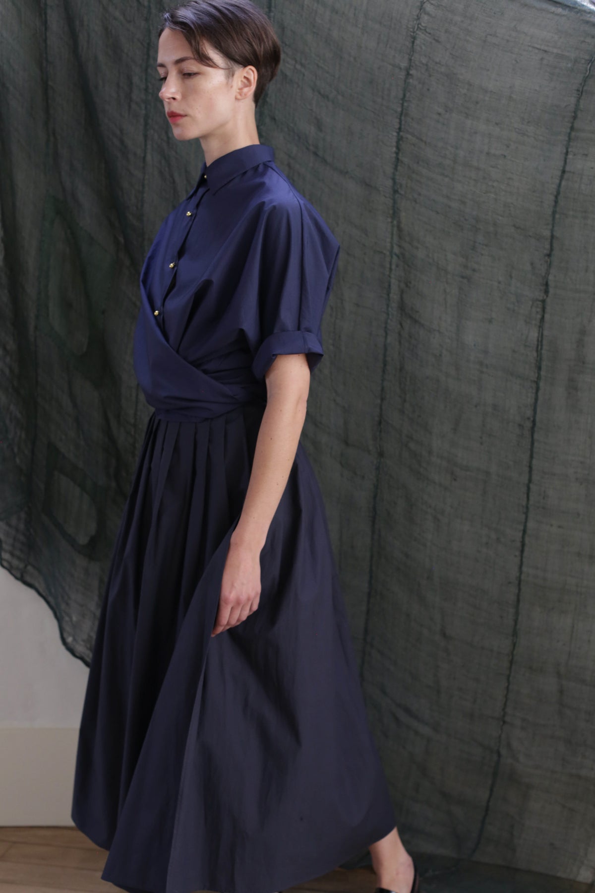 A person with short dark hair wears the Work Shirt in Typewriter Cotton Navy, standing in profile against a textured, dark green fabric backdrop.