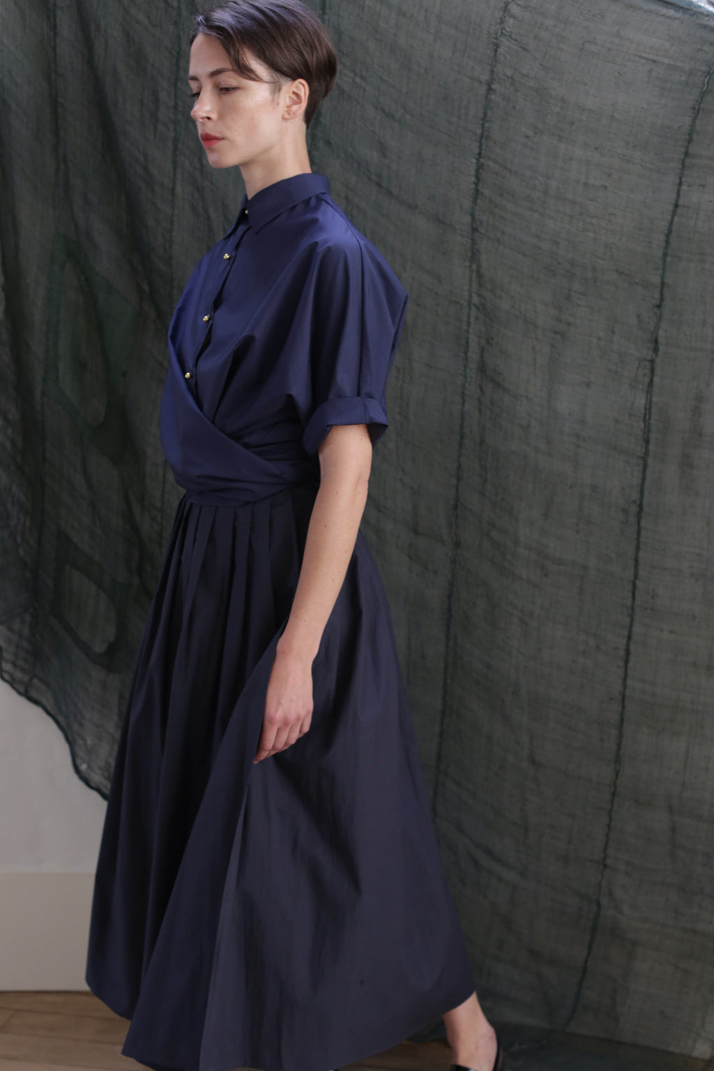 A person with short dark hair wears the navy Typewriter Wrap Skirt, standing in profile against a textured green fabric backdrop.