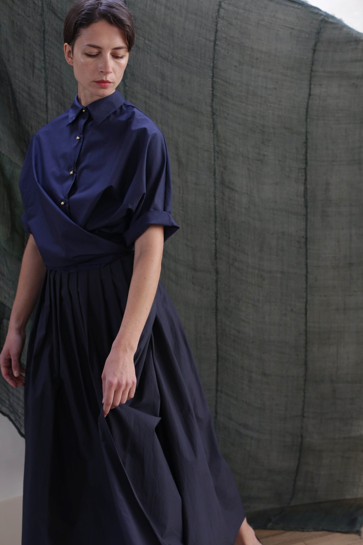 A woman wearing the Work Shirt | Typewriter Cotton Navy and a matching long skirt stands before a sheer dark green backdrop, looking down while gently holding her skirt.