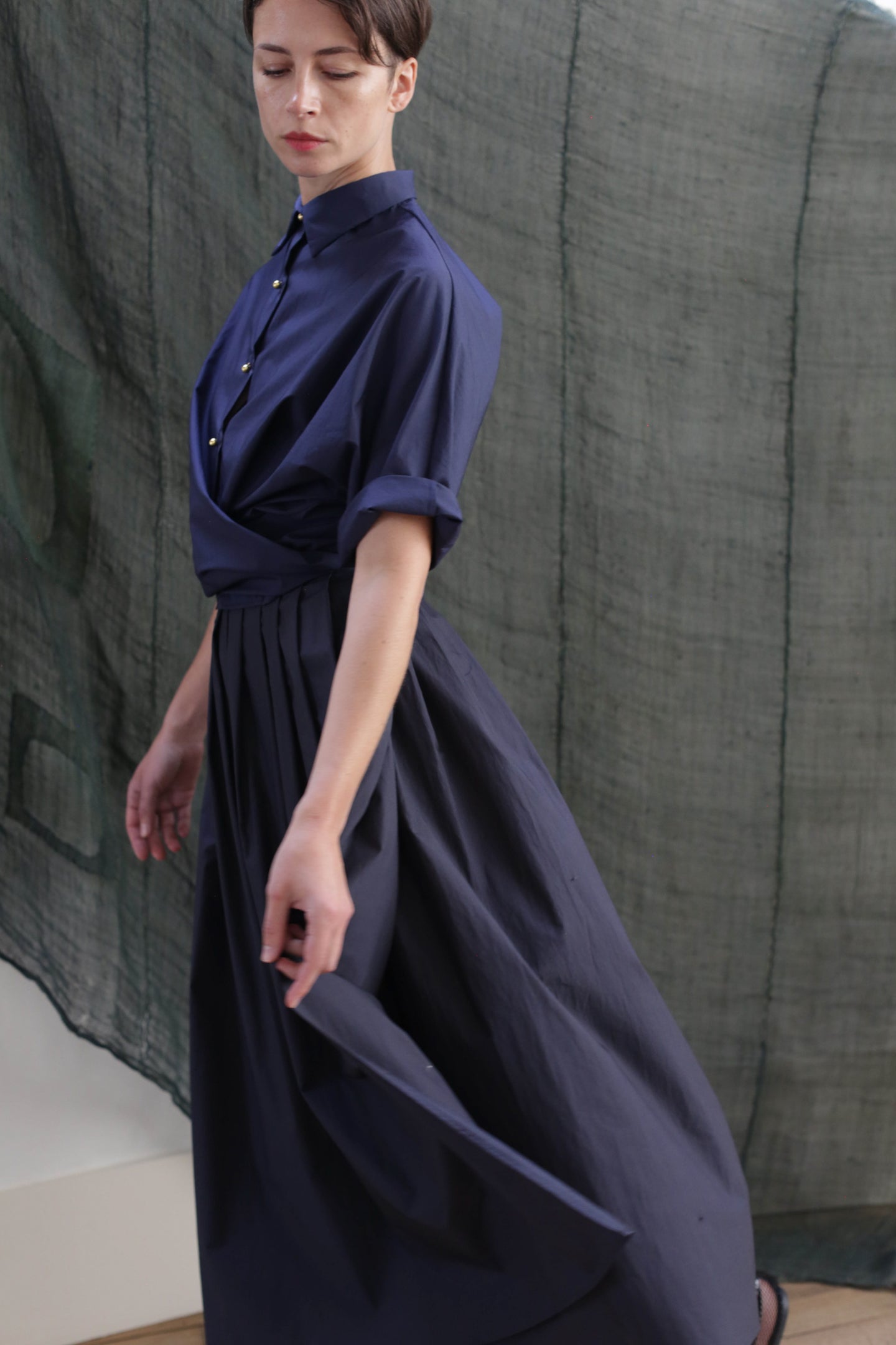 A woman wearing the Typewriter Wrap Skirt in navy, made from cotton and featuring elegant pleats, stands gracefully with one arm extended before a draped, semi-transparent green backdrop.