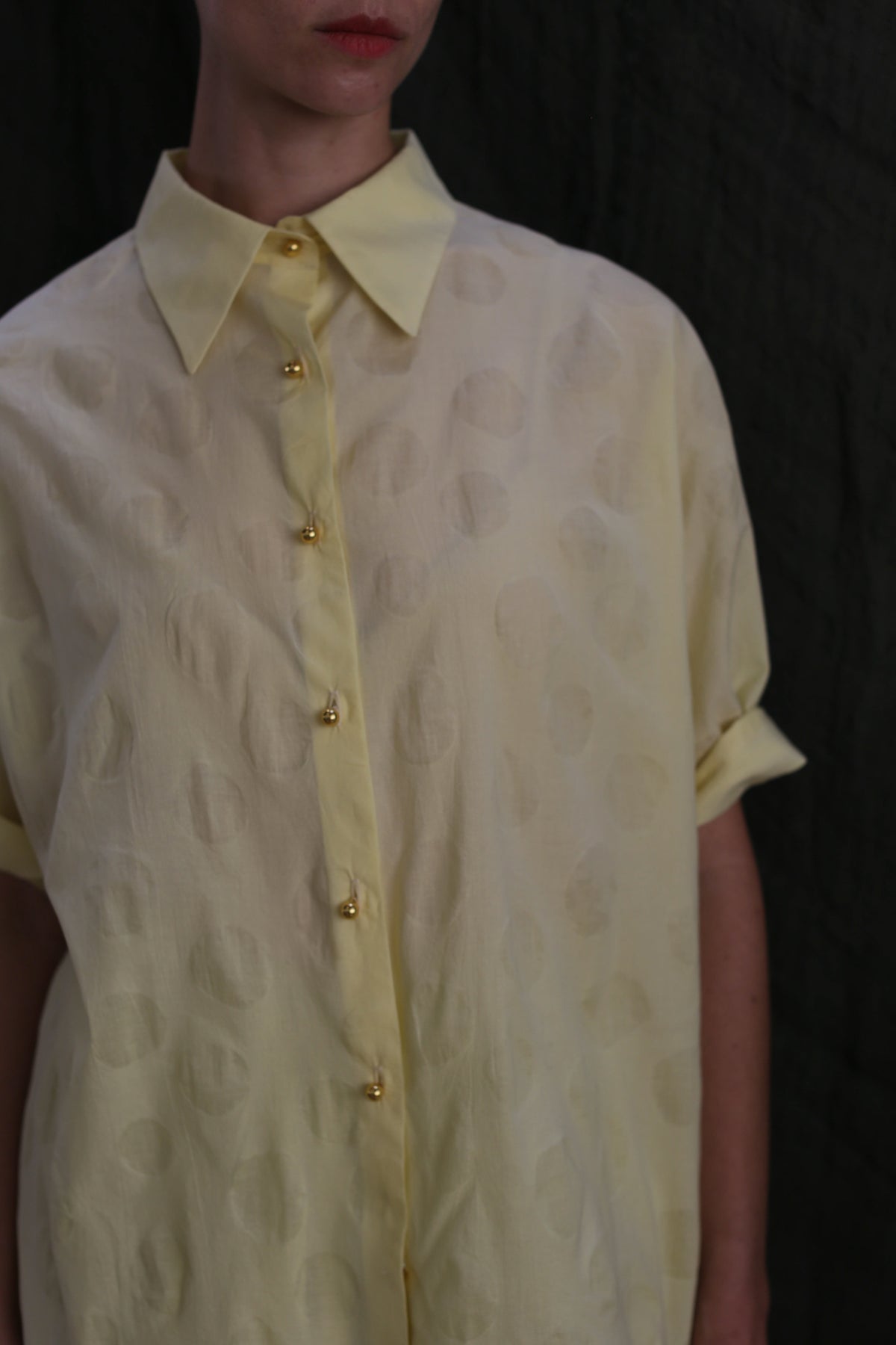 A person wears the Work Shirt | Textured Lawn Citrine—a pale yellow, short-sleeved cotton lawn shirt with subtle polka dots and brass buttons—standing against a dark background, their face partially out of frame.