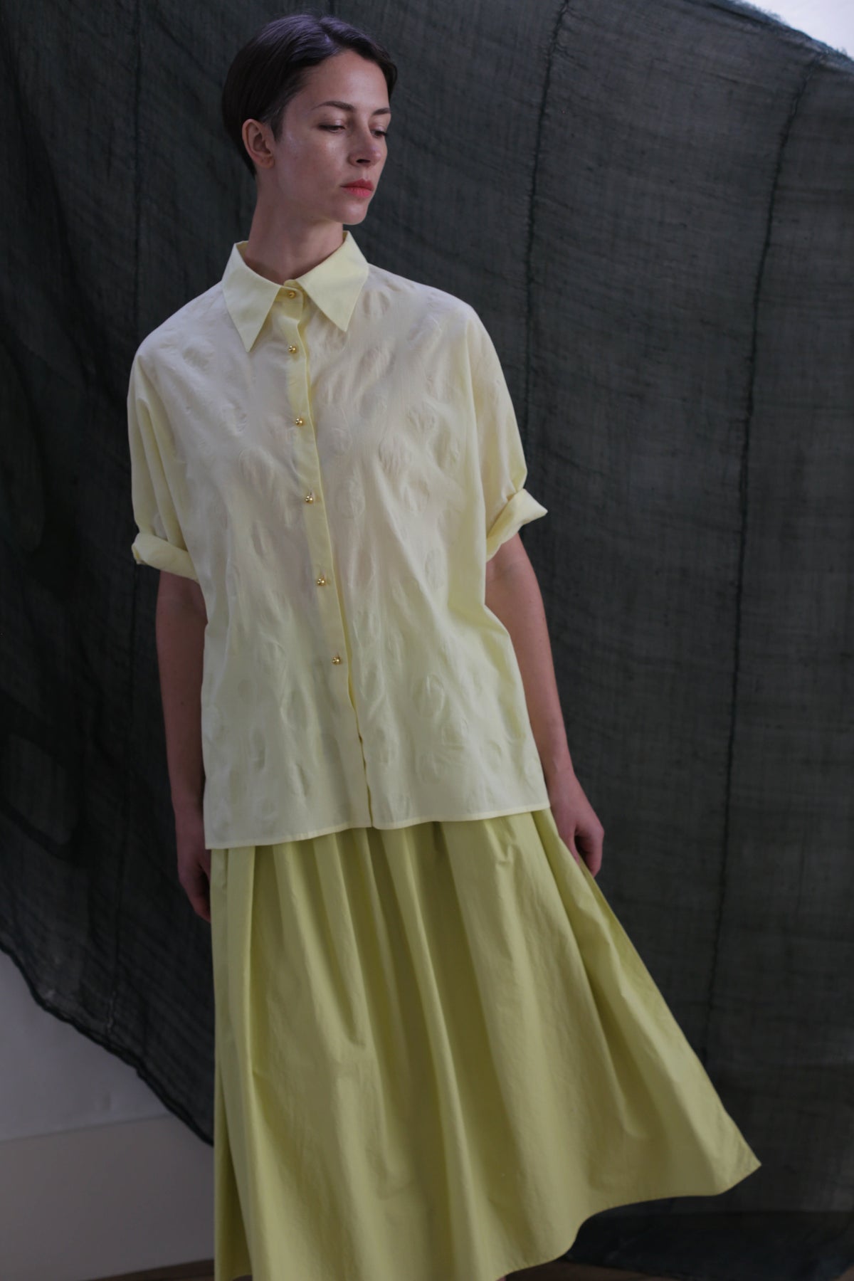 Against a dark, textured backdrop, a person wears the Work Shirt | Textured Lawn Citrine—a light yellow short-sleeve cotton lawn button-up with brass buttons—paired with a matching flowing skirt, head slightly tilted as they look to the side.