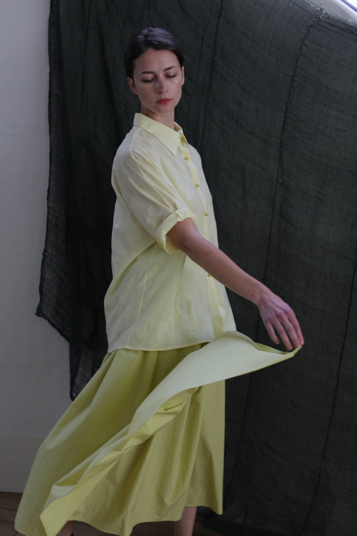 A person models the Work Shirt | Textured Lawn Citrine, paired with a matching flowing skirt, posing gracefully before a dark, sheer fabric backdrop.