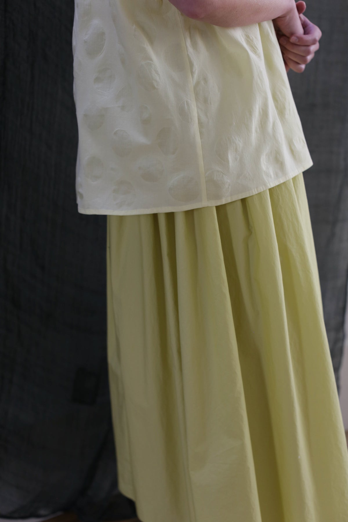 A person wears the Work Shirt in Textured Lawn Citrine with a matching flowing yellow skirt, standing in front of a dark fabric backdrop and clasping their hands behind their back.