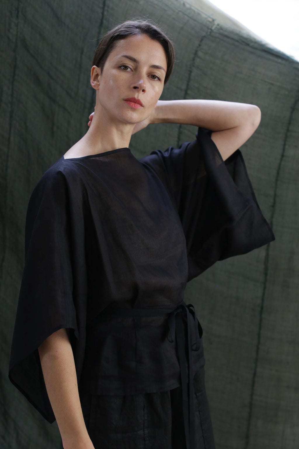 A woman wearing the Kimono Top | Ramie Black with wide sleeves stands before a dark green backdrop, gazing at the camera, one hand on her neck and the other by her side.