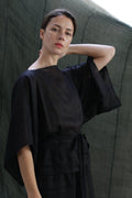 A woman wearing the Kimono Top | Ramie Black with wide sleeves stands before a dark green backdrop, gazing at the camera, one hand on her neck and the other by her side.