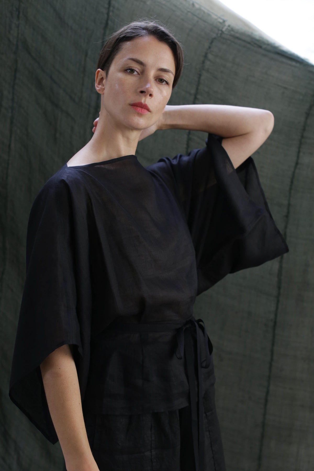 A woman wearing the Kimono Top | Ramie Black with wide sleeves stands before a dark green backdrop, gazing at the camera, one hand on her neck and the other by her side.