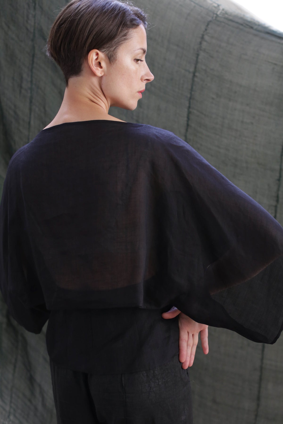 A person with short brown hair stands sideways, looking left. They wear the Kimono Top | Ramie Black, featuring wide sleeves and sheer black ramie fabric. One hand rests on their lower back against a muted green fabric backdrop.