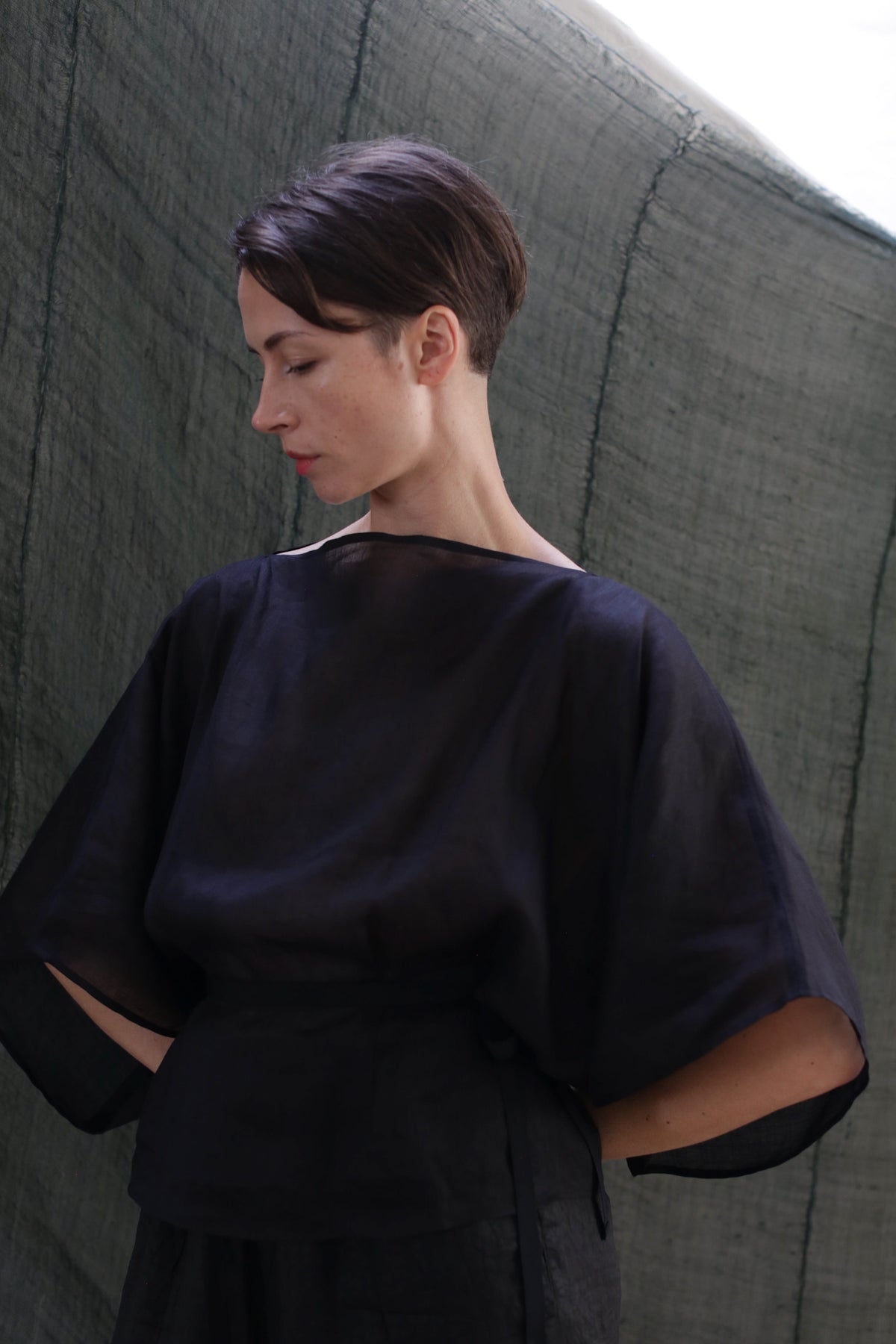 A person with short dark hair wears the Kimono Top | Ramie Black and stands against a textured dark green background, gazing downward with a calm, thoughtful expression.