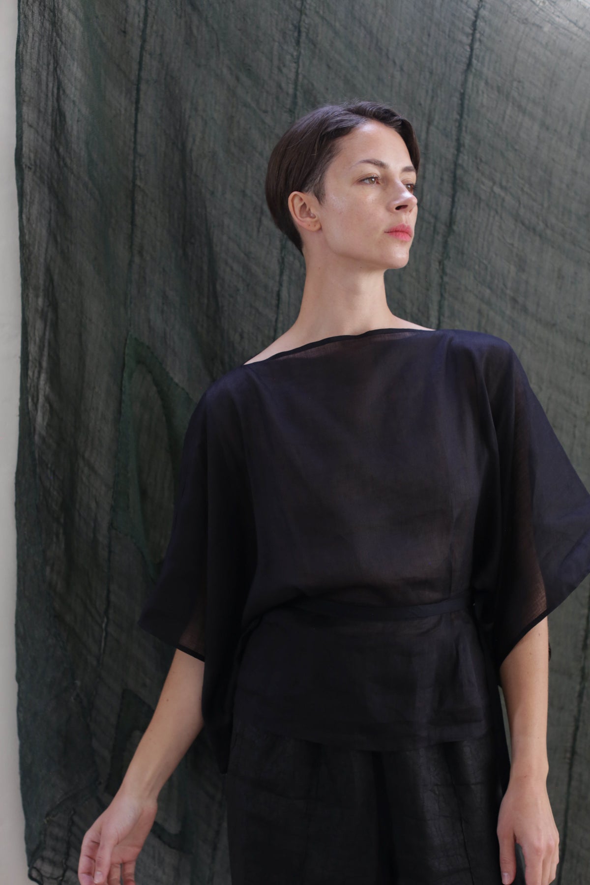 A woman with short brown hair wears the Kimono Top | Ramie Black, a loose, semi-sheer black top, and stands before a textured dark green backdrop, gazing slightly upward with a calm expression.