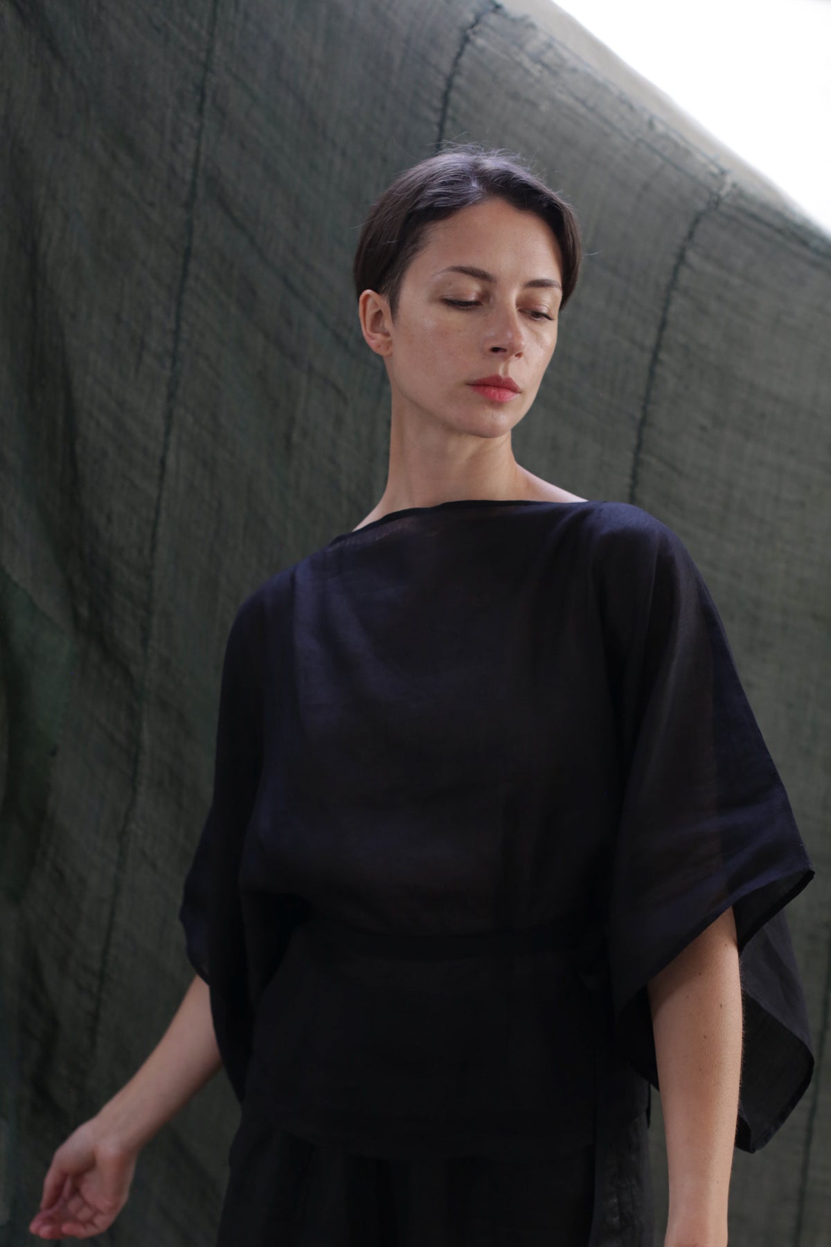 A woman with short dark hair stands before a textured green backdrop, wearing the Kimono Top | Ramie Black. The semi-sheer fabric drapes loosely as she looks downward with a calm expression, her face softly lit.