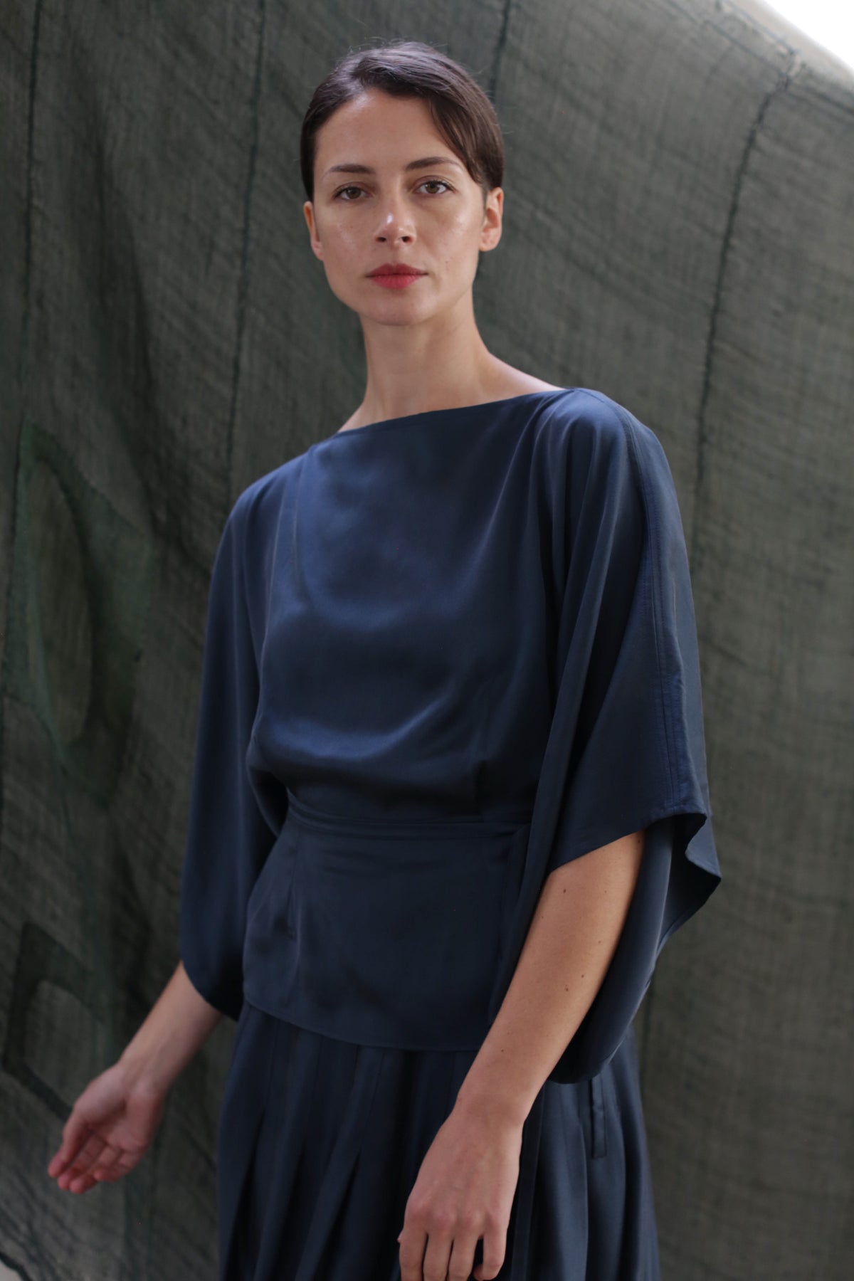 A woman with dark hair in a low bun models the Kimono Top | Sandwashed Silk Charmeuse Teal, featuring wide sleeves, as she stands before a textured dark green backdrop and looks calmly at the camera.