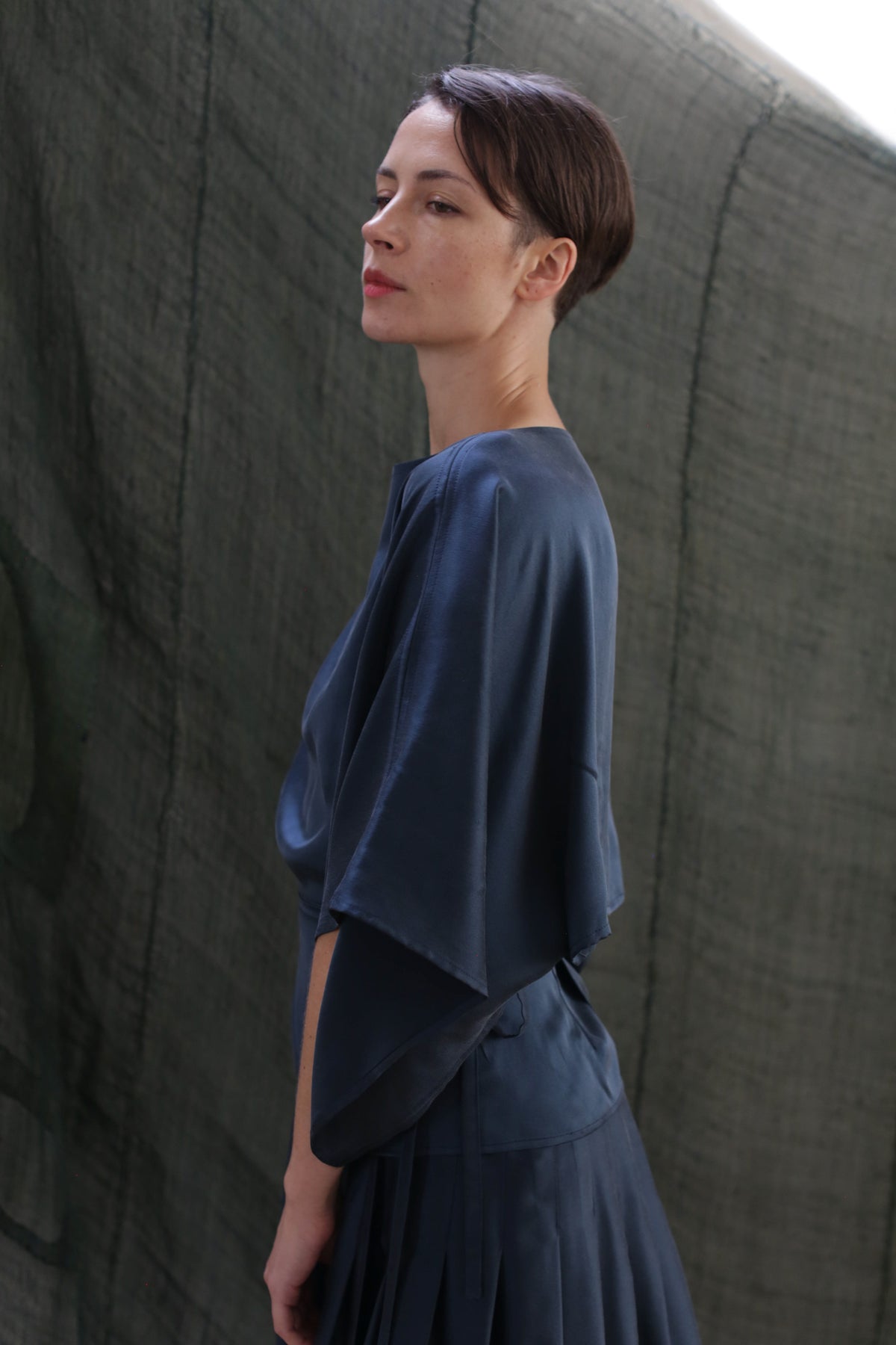 A woman with short brown hair wears the Kimono Top | Sandwashed Silk Charmeuse Teal, standing in profile against a muted green fabric backdrop and gazing upward with a calm expression.