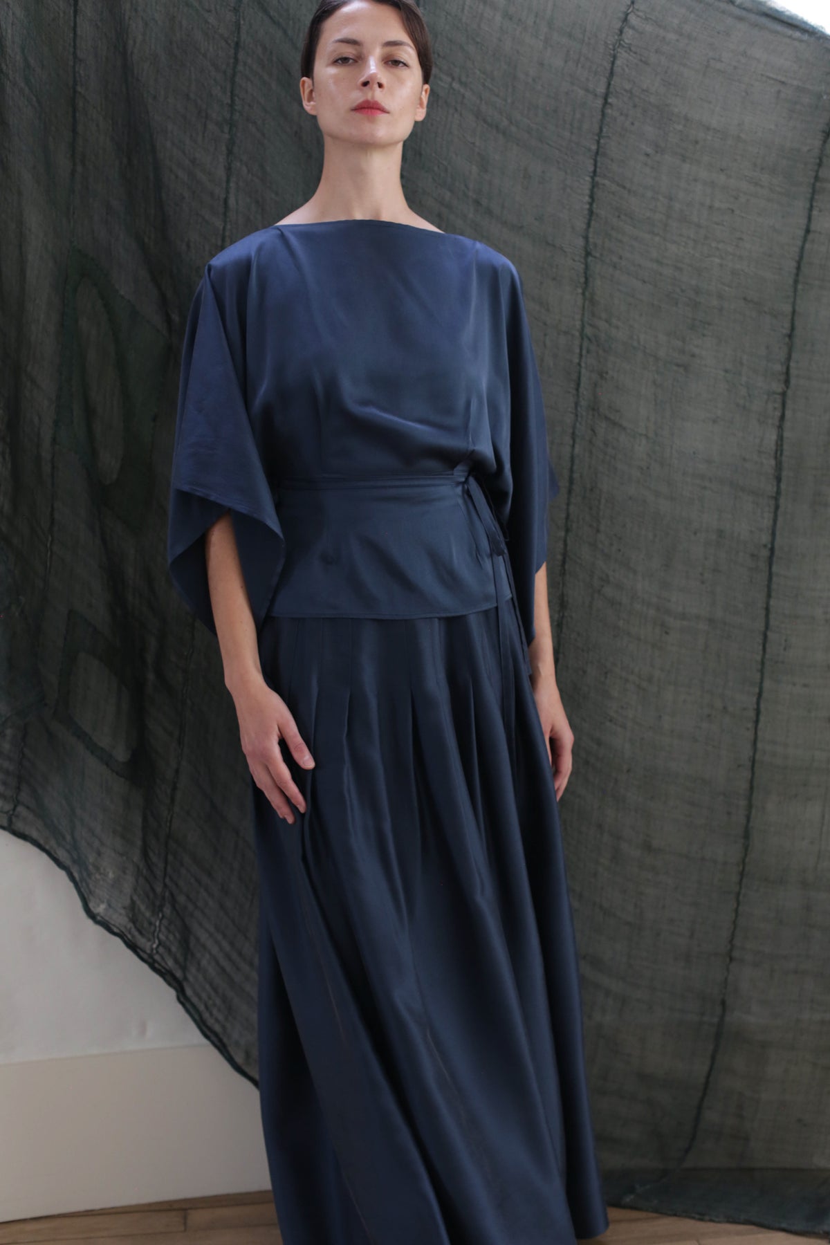 A woman stands indoors, calmly looking ahead while wearing the Hand Pleated Column Wrap Skirt in sandwashed silk charmeuse teal; wide sleeves and a cinched waist complete her look, set against a sheer textured dark backdrop.