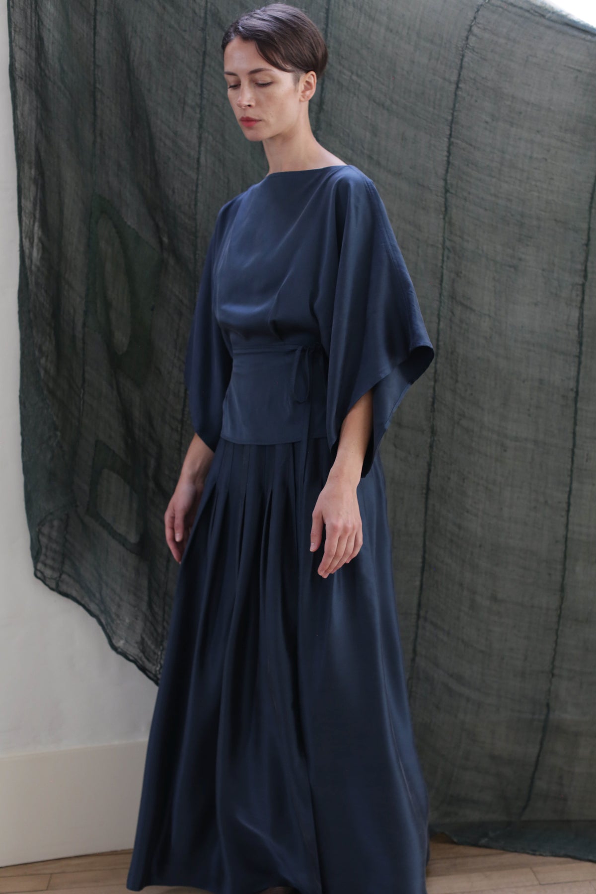 A woman stands indoors wearing the Hand Pleated Column Wrap Skirt in Sandwashed Silk Charmeuse Teal, paired with a flowing kimono-style top. In the background, sheer dark green fabric is draped against a light wall.
