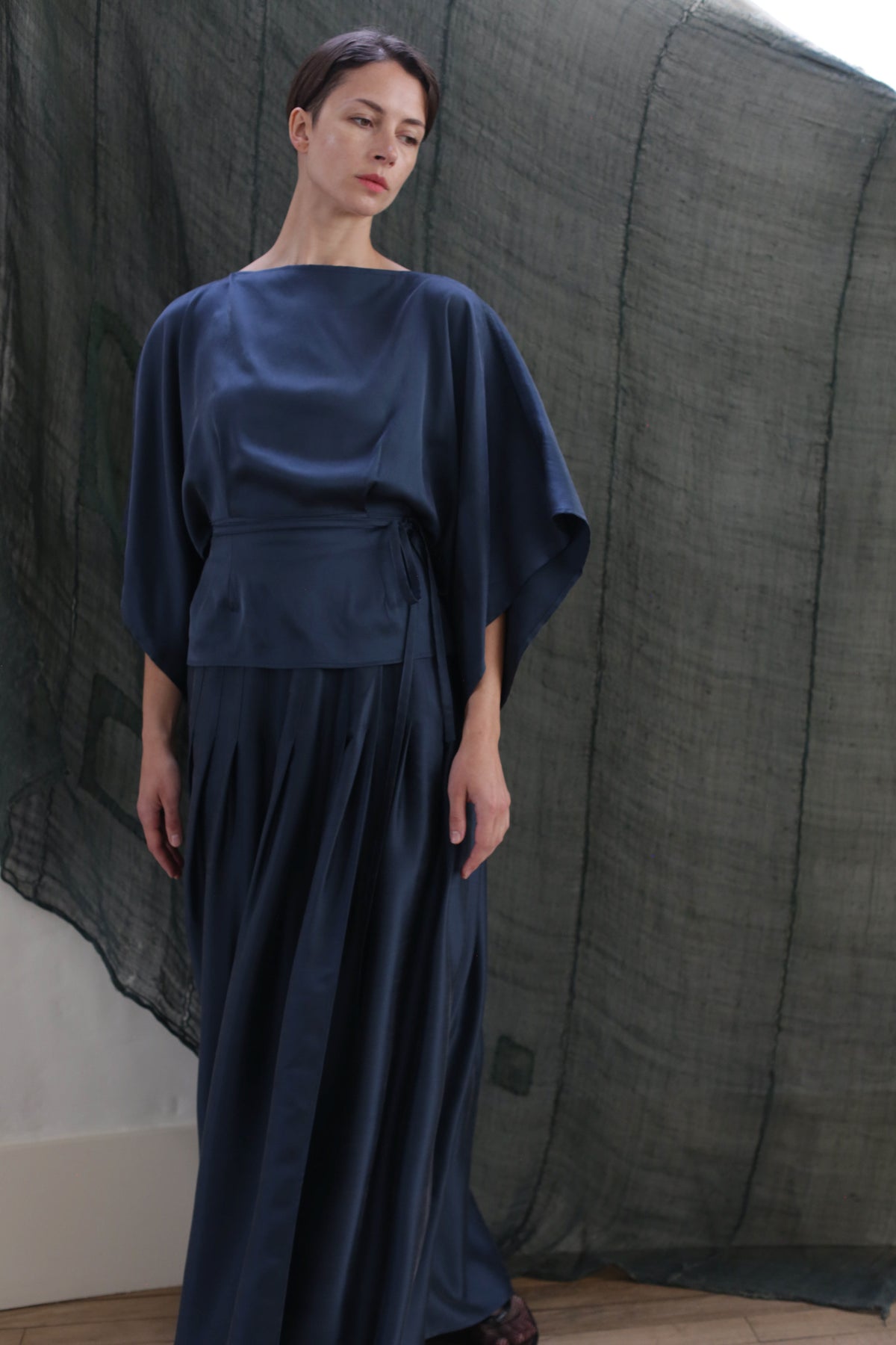 A woman wearing the Hand Pleated Column Wrap Skirt in sandwashed silk charmeuse teal stands before a sheer, dark green textured backdrop. She has short brown hair and gazes slightly downward with a neutral expression.