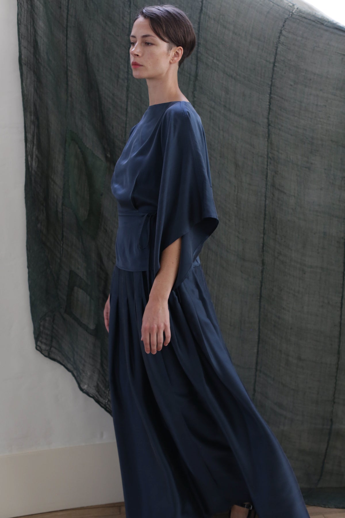 A woman with short dark hair stands in profile against a sheer dark green backdrop, wearing the Hand Pleated Column Wrap Skirt in sandwashed silk charmeuse teal with wide sleeves.