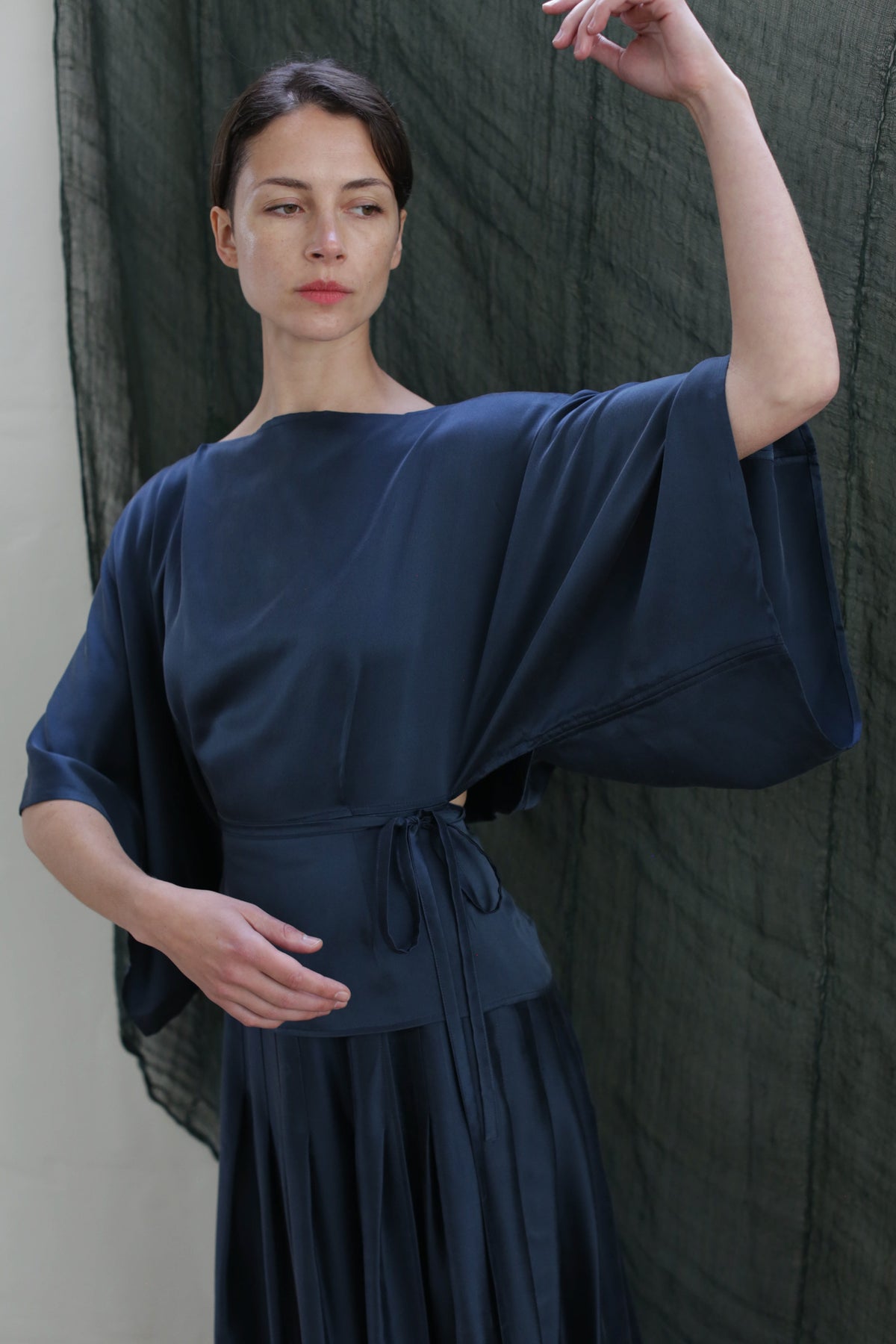 A woman in a flowing Kimono Top made of Sandwashed Silk Charmeuse in teal with wide sleeves stands before a dark textured backdrop, raising one arm and gazing slightly to the side.