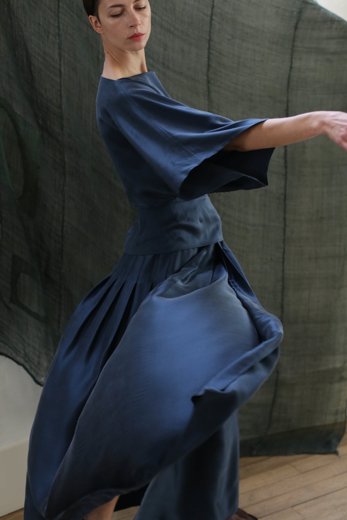 A person moves gracefully in the Kimono Top | Sandwashed Silk Charmeuse Teal, arms slightly extended as the teal fabric flows. The dark, textured background adds depth to this elegant zero-waste fashion scene.