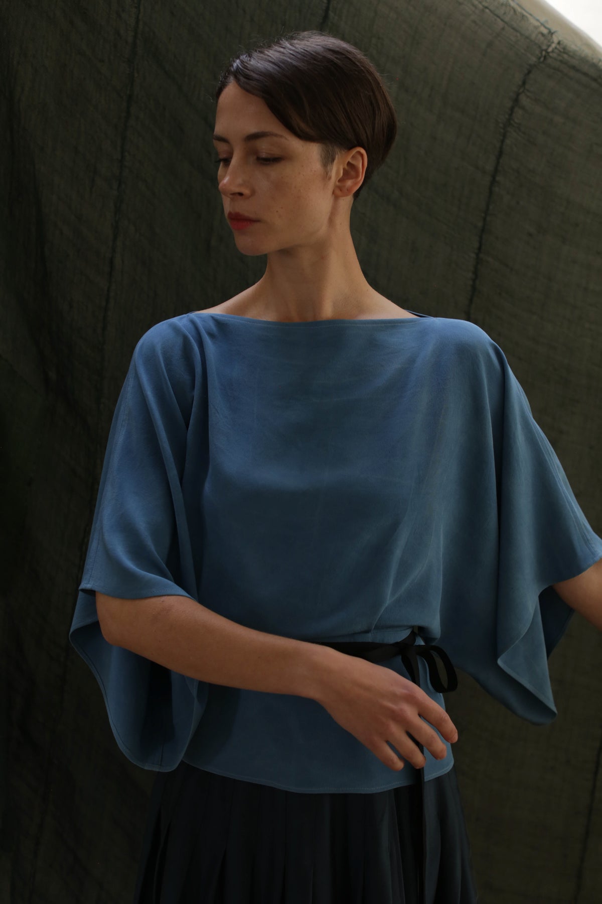 A woman with short dark hair stands against a dark, textured background wearing the Kimono Top | 2-ply Silk Crepe Ai Dye in blue, its rich hue suggesting an indigo-dyeing process. She looks down calmly in the loose-fitting style.