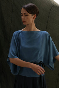 A woman with short dark hair stands against a dark, textured background wearing the Kimono Top | 2-ply Silk Crepe Ai Dye in blue, its rich hue suggesting an indigo-dyeing process. She looks down calmly in the loose-fitting style.