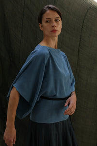 A woman with dark hair pulled back wears the Kimono Top | 2-ply Silk Crepe Ai Dye, featuring wide sleeves and a black belt, standing before a textured dark green backdrop. She gazes calmly to the side.