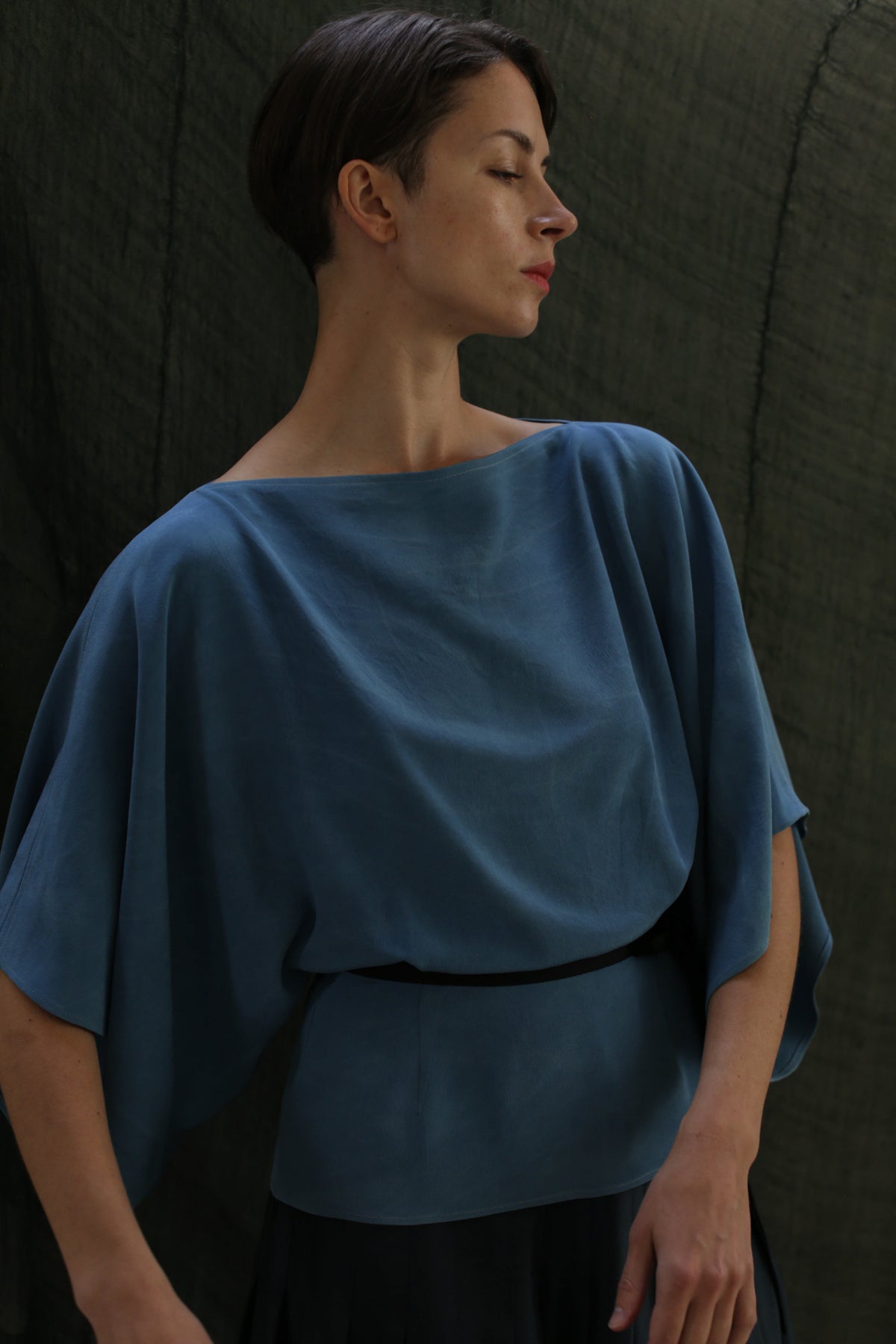 A person with short dark hair wears the Kimono Top | 2-ply Silk Crepe Ai Dye in blue, standing against a dark, textured background and gazing calmly to the side.