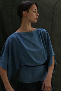 A person with short dark hair wears the Kimono Top | 2-ply Silk Crepe Ai Dye in blue, standing against a dark, textured background and gazing calmly to the side.