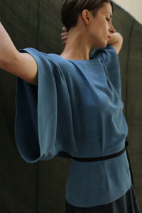 A person poses with arms raised and head turned, wearing the Kimono Top | 2-ply Silk Crepe Ai Dye in blue against a dark, textured background.