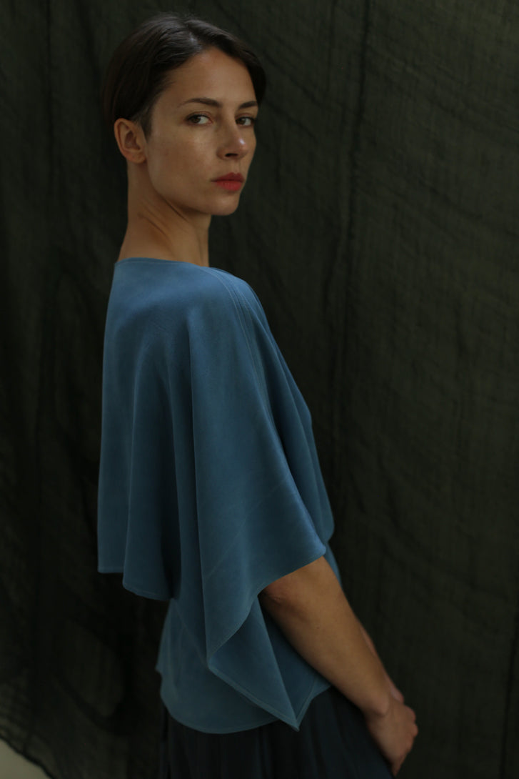 A person with short dark hair and red lipstick wears a loose-fitting Kimono Top | 2-ply Silk Crepe Ai Dye, standing against a dark textured background and glancing over their shoulder at the camera.