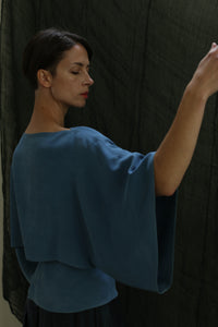A woman with short dark hair wears a loose Kimono Top | 2-ply Silk Crepe Ai Dye, standing before a dark textured background, her eyes closed and one arm gracefully extended to the side.