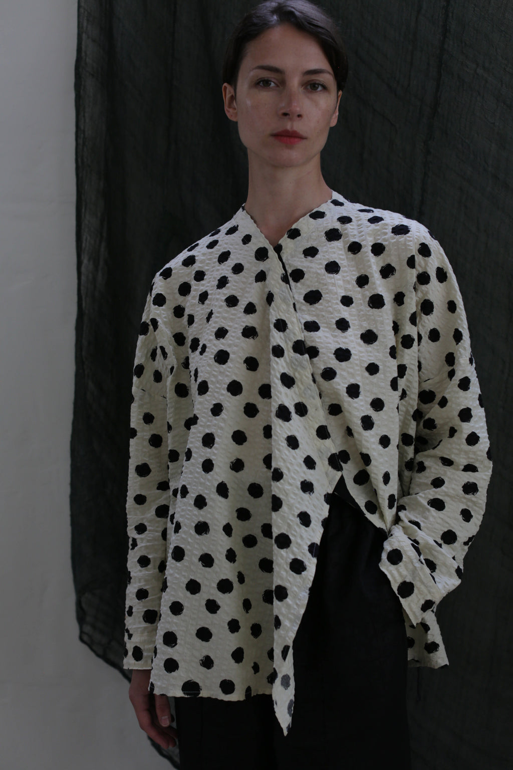 A person poses against a dark textured background, wearing the Qipao Blouse | Polkadot Seersucker Ivory with black pants and one hand in their pocket.