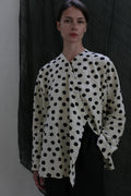 A person poses against a dark textured background, wearing the Qipao Blouse | Polkadot Seersucker Ivory with black pants and one hand in their pocket.