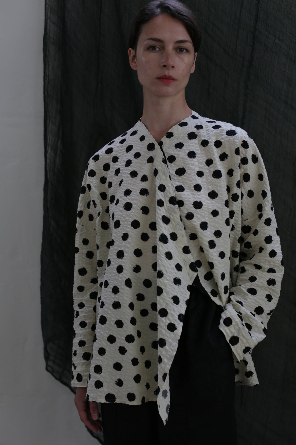 A person poses against a dark textured background, wearing the Qipao Blouse | Polkadot Seersucker Ivory with black pants and one hand in their pocket.