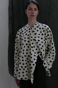 A person poses against a dark textured background, wearing the Qipao Blouse | Polkadot Seersucker Ivory with black pants and one hand in their pocket.