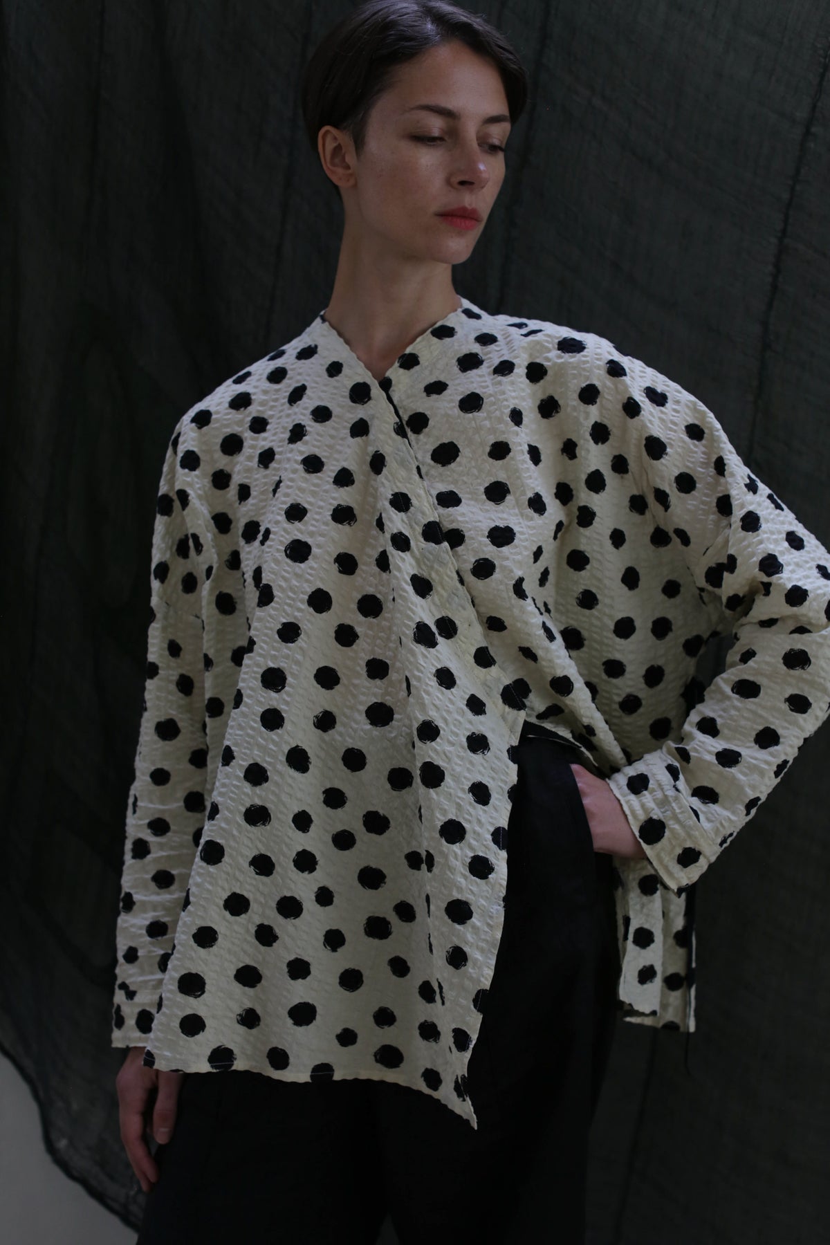 A person with short hair wears the Qipao Blouse | Polkadot Seersucker Ivory and black pants, standing against a dark draped background. Their hand rests on their hip as they look slightly downward.