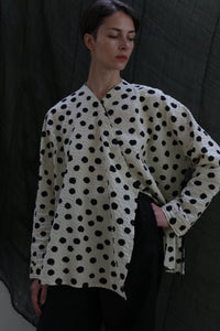 A person with short hair wears the Qipao Blouse | Polkadot Seersucker Ivory and black pants, standing against a dark draped background. Their hand rests on their hip as they look slightly downward.