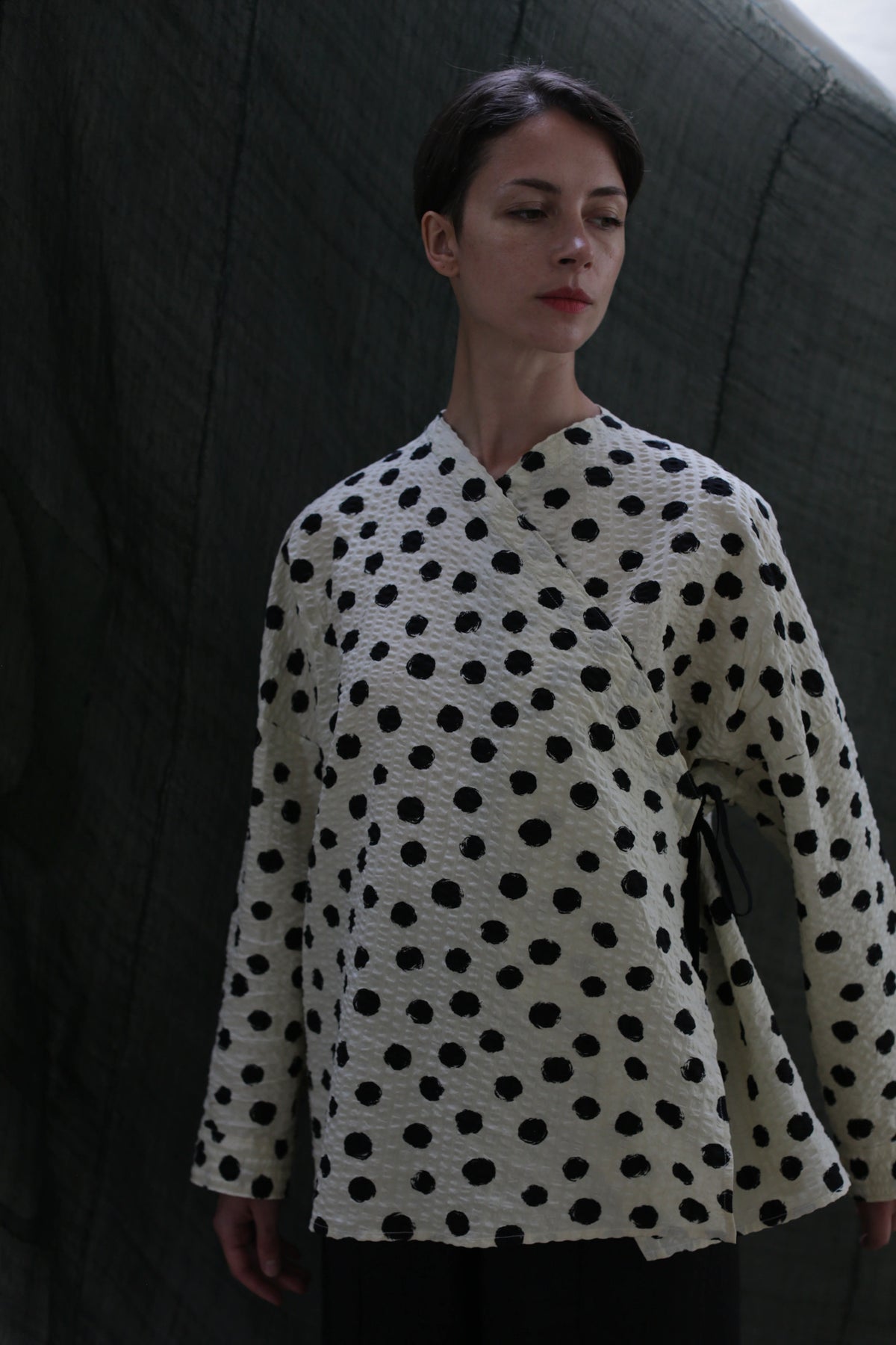 A woman with short dark hair wears the Qipao Blouse | Polkadot Seersucker Ivory, standing before a dark fabric backdrop. She looks down calmly, highlighting the blouse’s cream color and black polka dot pattern.