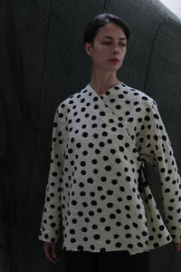 A woman with short dark hair wears the Qipao Blouse | Polkadot Seersucker Ivory, standing before a dark fabric backdrop. She looks down calmly, highlighting the blouse’s cream color and black polka dot pattern.