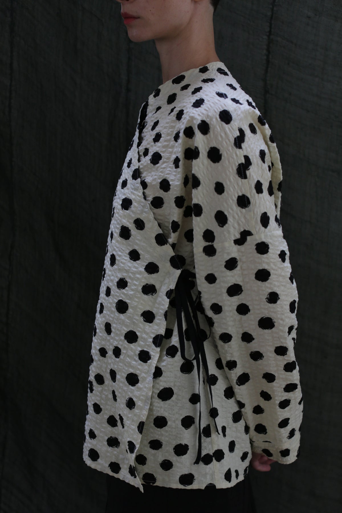 A person stands sideways wearing the Qipao Blouse | Polkadot Seersucker Ivory—a cream wrap top with black polkadots and side ribbon ties—against a dark, textured fabric background.