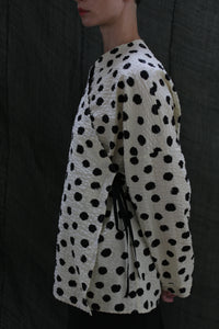 A person stands sideways wearing the Qipao Blouse | Polkadot Seersucker Ivory—a cream wrap top with black polkadots and side ribbon ties—against a dark, textured fabric background.