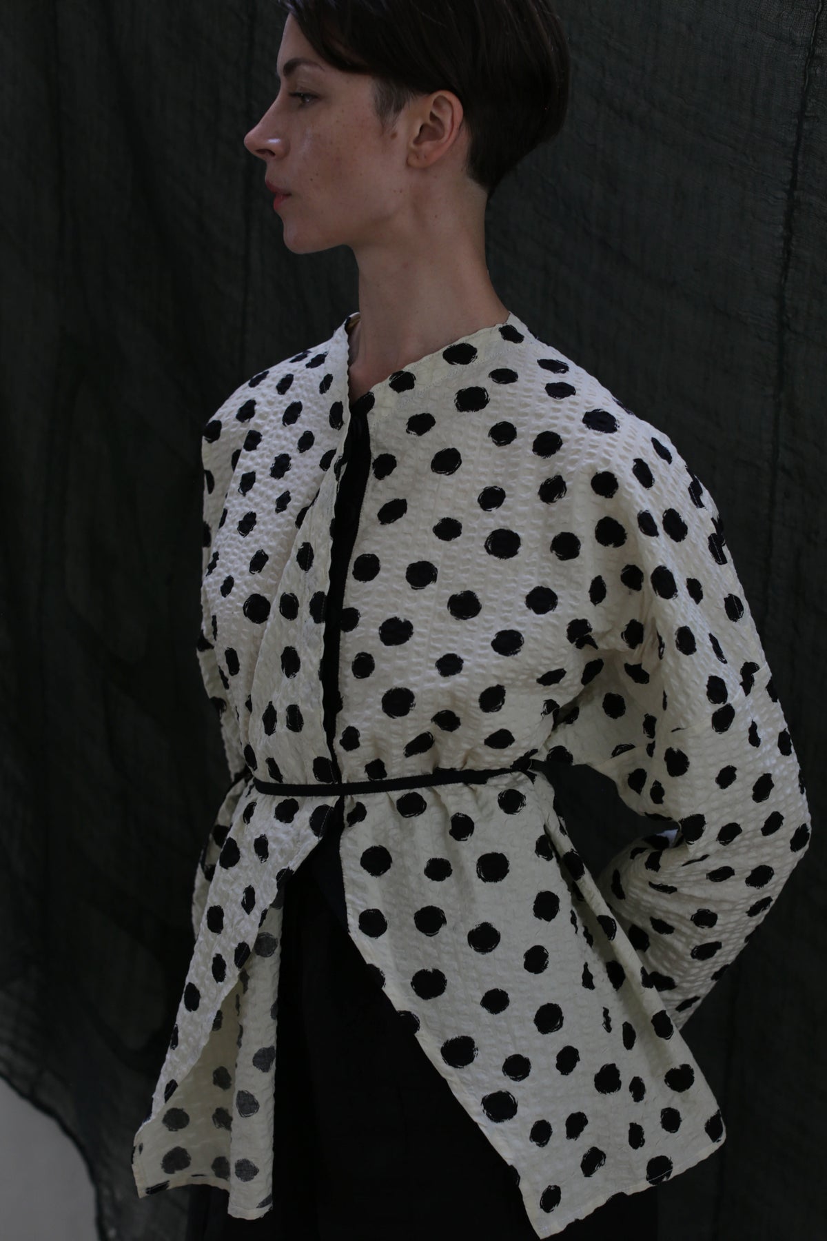 A person with short dark hair models the Qipao Blouse | Polkadot Seersucker Ivory, featuring large black polkadots and a thin black waist belt, standing in profile before a dark, draped backdrop.