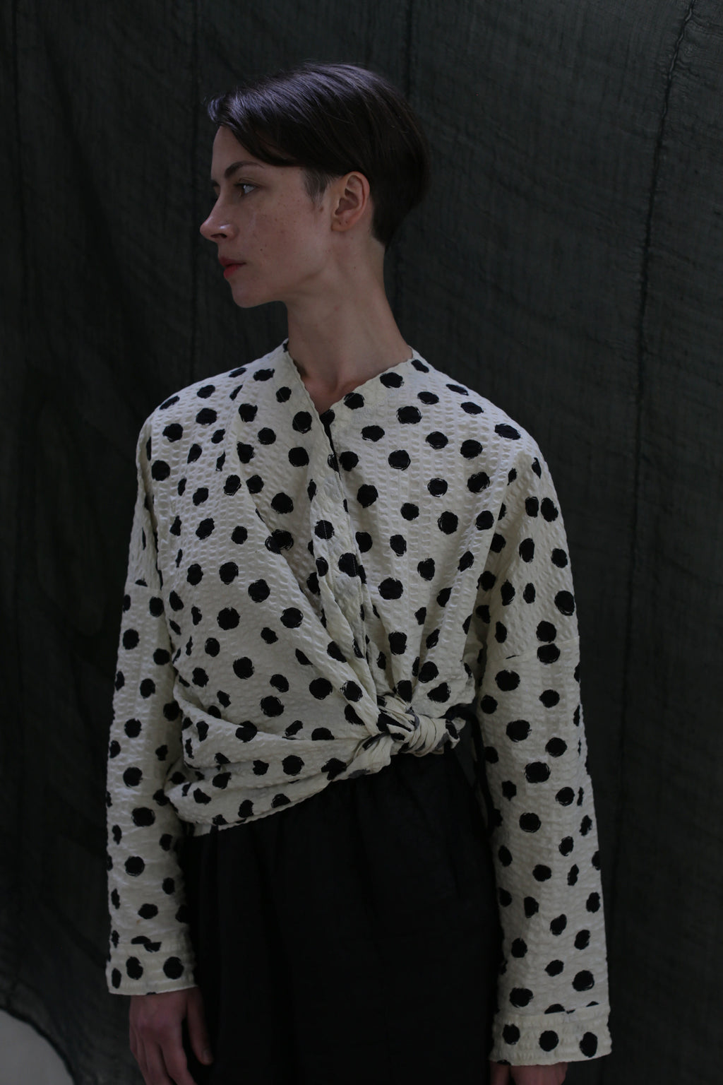 A person with short brown hair wears the Qipao Blouse | Polkadot Seersucker Ivory, styled with black pants, standing against a dark textured backdrop and looking to the side.