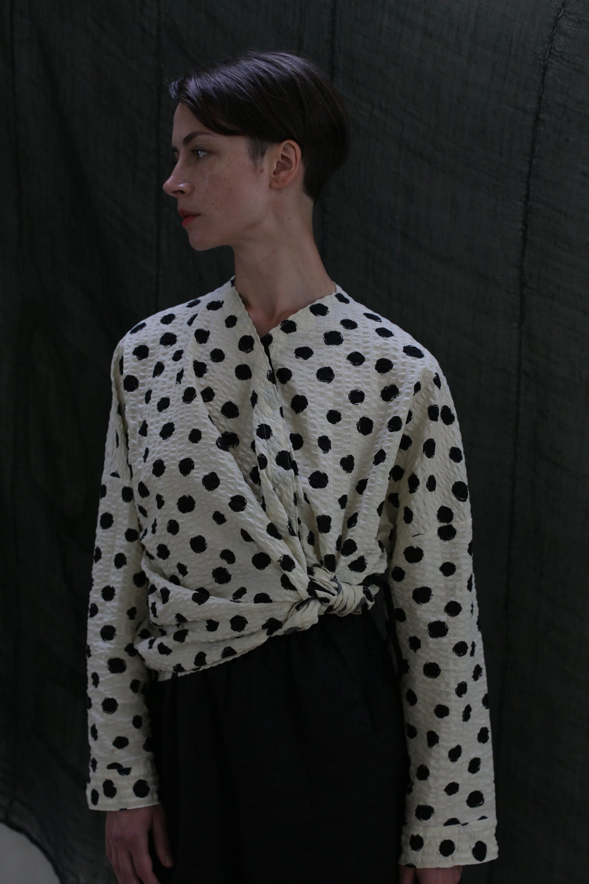 A person with short brown hair wears the Qipao Blouse | Polkadot Seersucker Ivory, styled with black pants, standing against a dark textured backdrop and looking to the side.