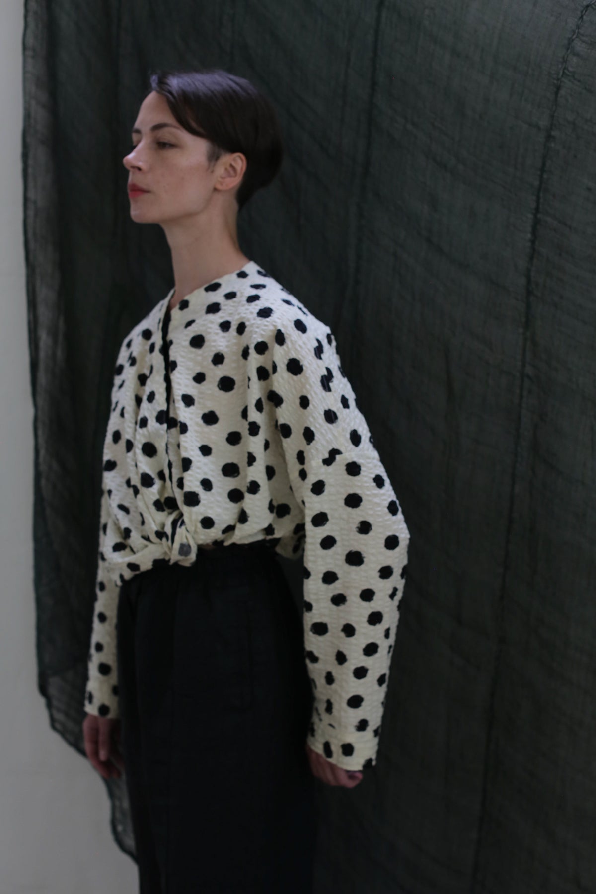 A person with short hair wears the Qipao Blouse | Polkadot Seersucker Ivory, styled with high-waisted dark pants, standing before a draped dark fabric background.