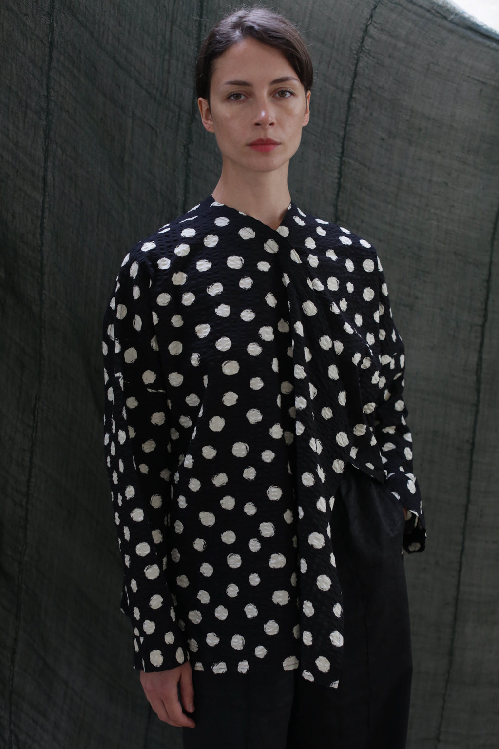 A person with dark hair pulled back wears the Qipao Blouse | Polkadot Seersucker Black, paired with black pants, standing before a dark textured background.